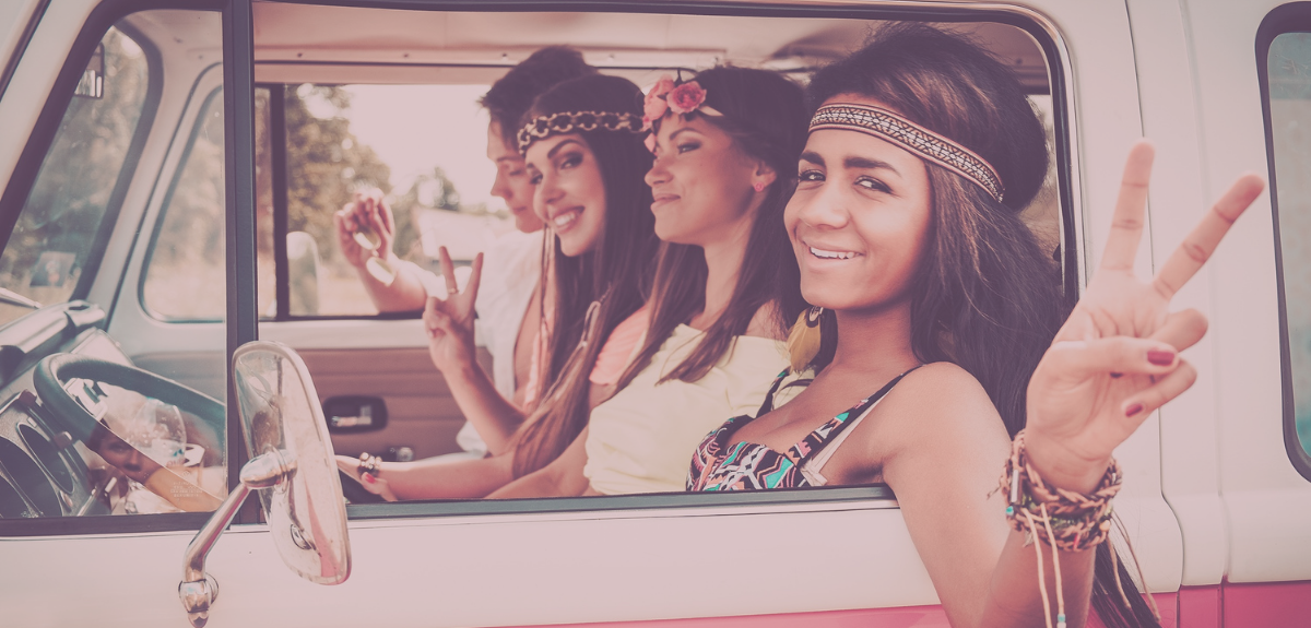 Four female friends sit in a retro bus while the one closest to the camera holds up a peace sign.